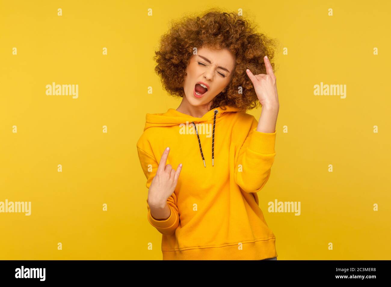 Yeah, that`s wonderful! Portrait of overjoyed curly-haired woman in urban style hoodie showing rock and roll hand sign, screaming and gesturing to hea Stock Photo